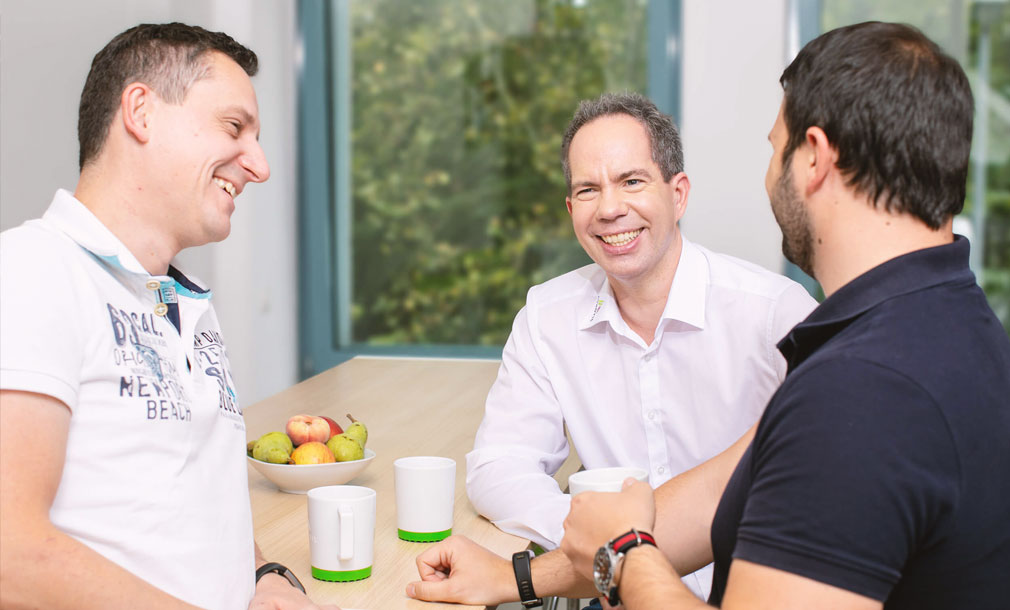 Drei lächelnde Personen sitzen an einem Tisch und unterhalten sich entspannt bei einer Tasse Kaffee. Auf dem Tisch stehen weiße Tassen und eine Schale mit Obst.