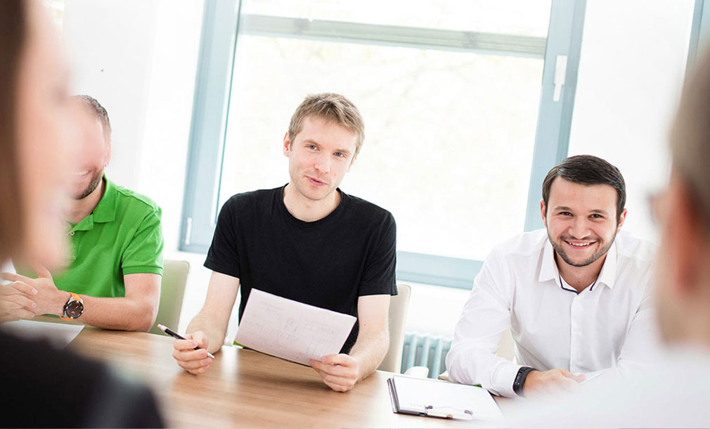 Personen sitzen an einem Konferenztisch und diskutieren, eine Person hält Notizen in der Hand. Zwei von ihnen lächeln in die Kamera.